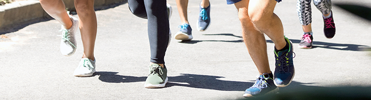 Lower part of legs and feet of multiple runners on pavement in various athletic running shoes.