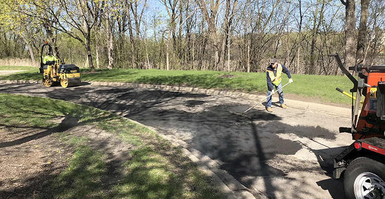 Two workers on curved roadway; one with handheld pusher smoothing freshly filled asphalt the other driving a heavy equipment rolling packer.