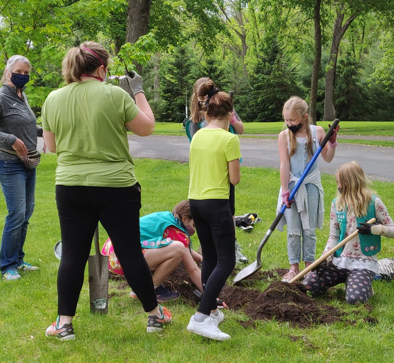 Group of five girls some with green Girl Scout vests with face coverings shoveling dirt to make large hole, two leaders supervising.