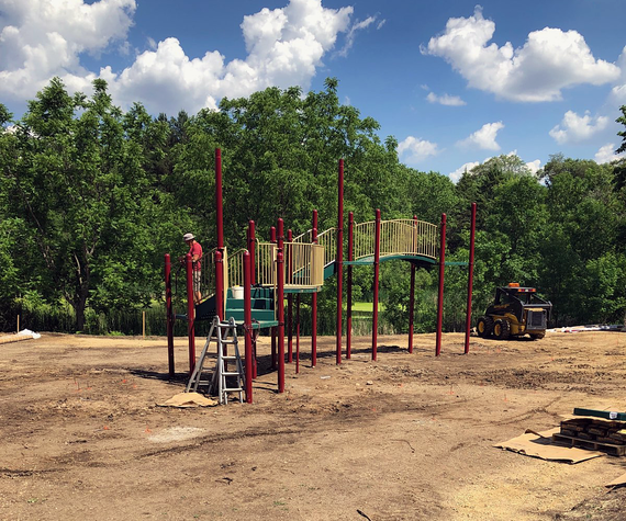 Playground structure forming with raised posts and elevated walkways, worker assembling structure in hard hat next to ladder over large dirt base.