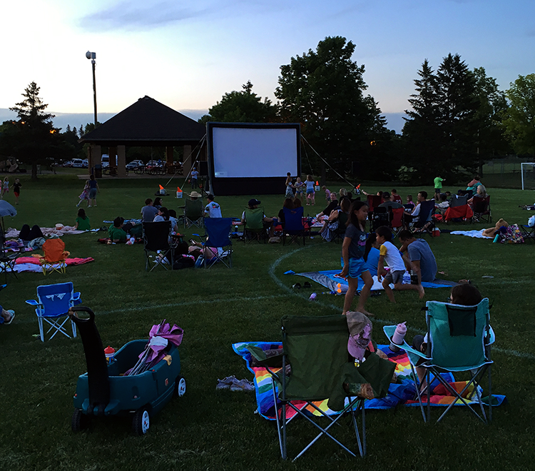 Inflatable movie screen in background surrounded by adults and kids in lawn chairs and on blankets facing screen at dusk.