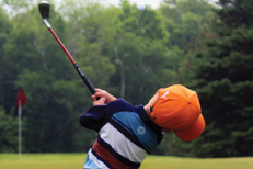 back of boy in orange hat and striped shirt swinging golf club above head towards green with flag