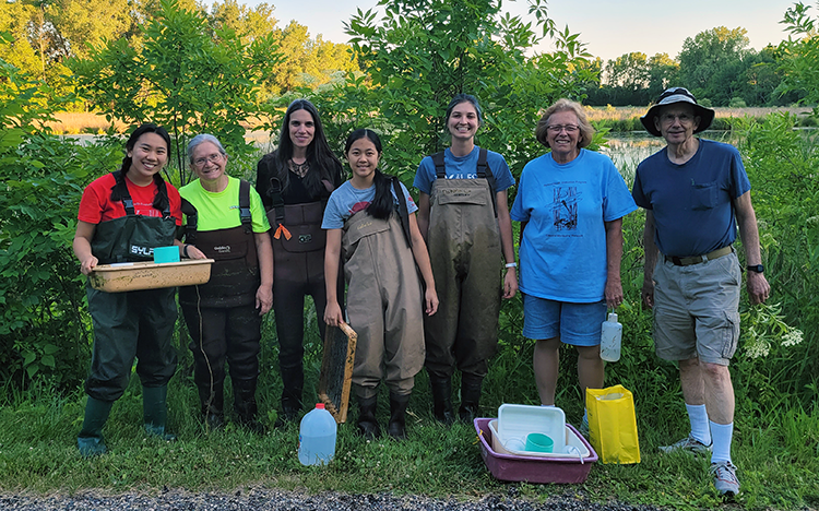 Group of seven smiling volunteers, several in bib waterproof waders, smiling in front of wetland, with bins and monitoring equipment.