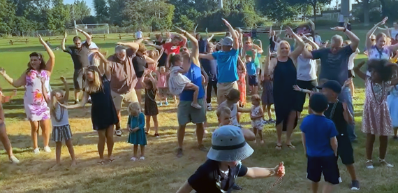 Crowd of adults and young kids with arms in the air dancing on park field.