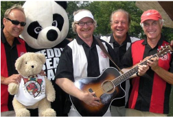 Group of four men smiling, one holding a guitar, one holding small stuffed teddy bear, with large stuffed panda bear in background.