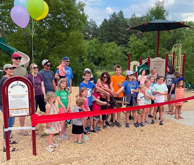 Group of children and adults in front of playground equipment with Mayor and kids holding gigantic scissors, cutting large red ribbon.
