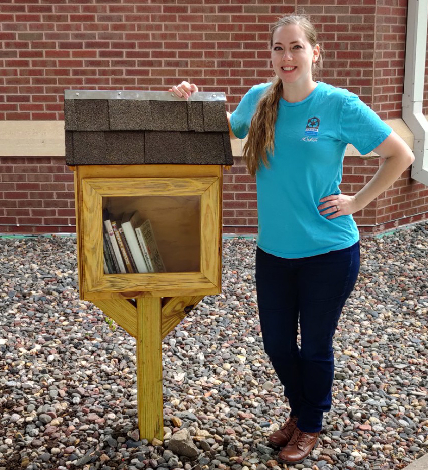 Woman in front of brick building smiling with hand on small, post-mounted wooden library, with books showing through clear window.