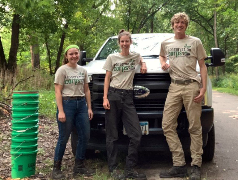 Three youth smiling in front of truck with tan Conservation Corps t-shirts, long pants and boots, tower of green buckets off to the side.