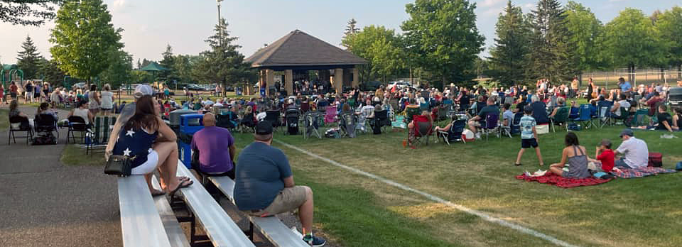 Large crowd of people facing the Mendakota Park pavilion; some sitting on bleachers and some on blankets and folding chairs.