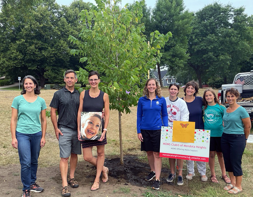 Group of people in front of newly planted tree, woman holding large photo of brightly smiling girl, Moms Club staked signage in front.