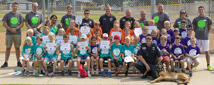 Large group of kids on bleachers with public safety personnel standing in back, uniformed officer kneeling with K9 dog in front.