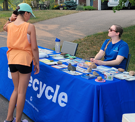 Woman sitting at Recycle table, printed materials being held down by rocks, youth in baseball cap browsing information. 