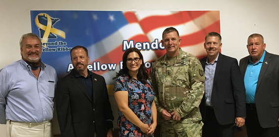 Group of five men and one woman smiling, with stars and stripes banner in the background featuring Beyond the Yellow Ribbon logo.