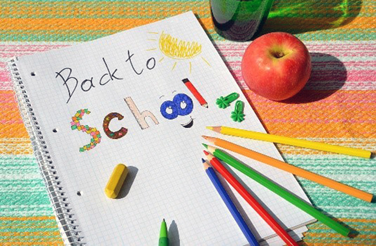 Spiral notebook with assorted colored pencils and hand-written text back-to-school over brightly colored and striped tablecloth with red apple.