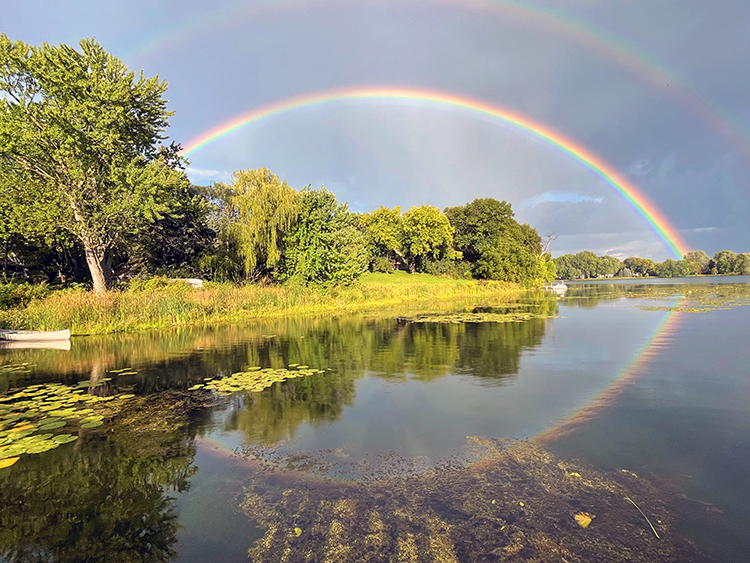 A large double rainbow spans the lake reflecting into the water in the foreground near green tree-lined shoreline.