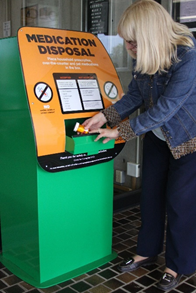 Woman standing and inserting prescription container into a large green metal drop box, front of box displays signage "Medication Disposal" 