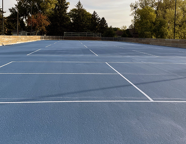 New blue surfaced pickleball courts with white lines, bordered on sides and back with rink boards, trees in background at Friendly Hills Park.