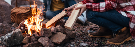 Person in red flannel shirt and boots kneeling next to outdoor bonfire tossing in two clean wood pieces.