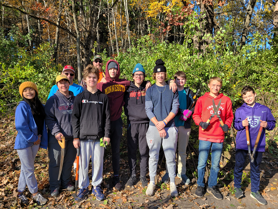Group of smiling youth dressed in workwear with adults in back, several holding clipping tools, against fall-colored tree background.