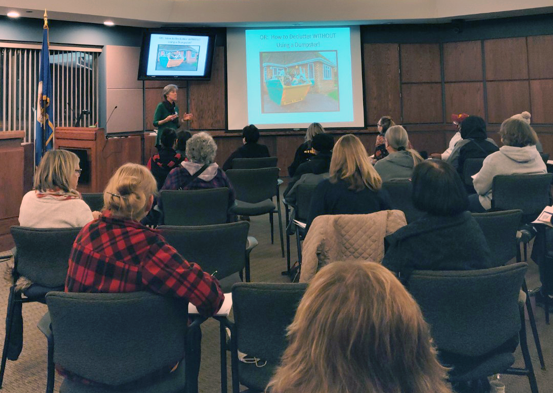 Back of seated attendees in council chambers with presenter at front, next to screen "How to declutter without using a dumpster"