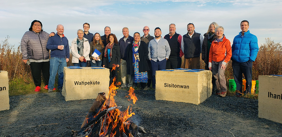 Group of sixteen standing behind large Council Fires stones imprinted with Dakota Names Wahpekute and Sisitonwan, bonfire in foreground.