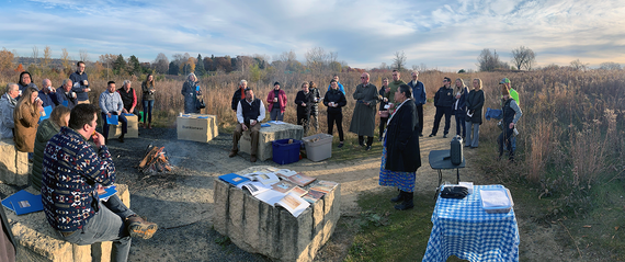 Group of people some sitting on large Seven Council Fire stones and some standing around bonfire near field, listening to speaker. 
