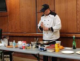 Chef standing over sauce pan behind table spread with various ingredients. 