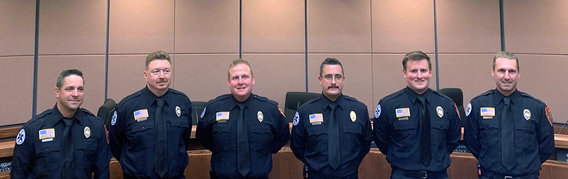 Six men in Mendota Heights firefighter dress uniforms with badges pinned smiling in City Hall Council Chambers.