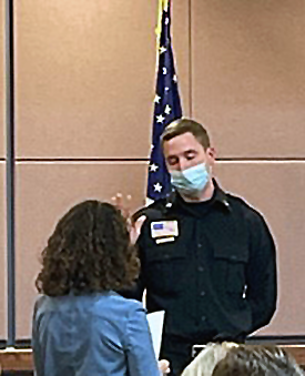 Man in Mendota Heights firefighter dress uniform holding up right hand in front of American flag, being sworn in by Mayor Levine.
