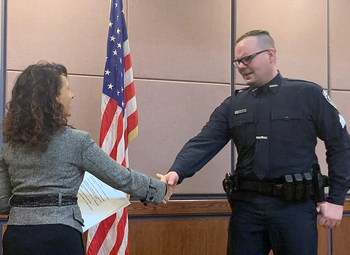 Officer in MHPD uniform shaking hands with Mayor Levine in Council Chambers after the swearing in, American Flag in background.
