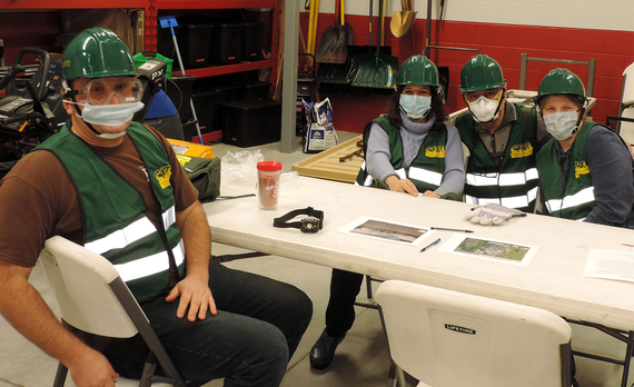 Four people sitting at a table at the fire station, dressed in CERT reflective safety vests wearing hard hats and face masks.