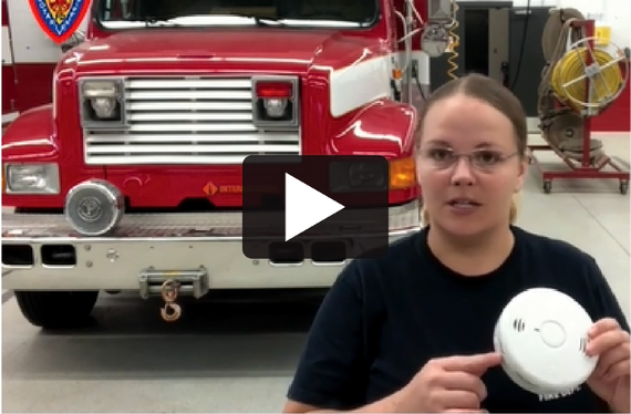 Firefighter holding up a CO detector with fire truck in background; icon to play video.