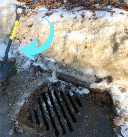 Cleared storm drain with arrow pointing to large metal shovel off to one side, surrounded by piled up snow and ice.