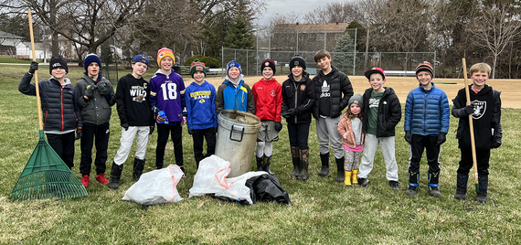 Group of smiling kids in coats and knit hats in front of ballfield, a couple with rakes, standing behind bags of garbage collected from the park.