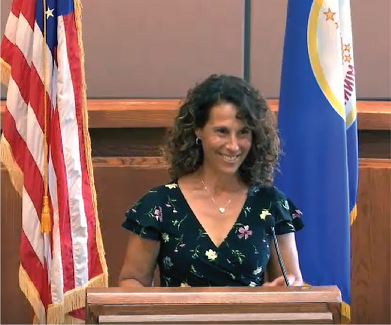 Mayor Levine smiling at the podium in the City Council Chambers with American flag and Minnesota State flag in background.