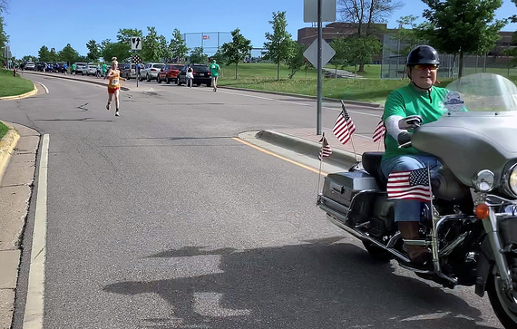 Man on motorcycle decorated with American flags leading the way with runners in background on roadway.