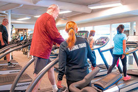 People exercise on treadmills in a gym, including an individual walking with assistance from someone wearing a 'Personal Trainer' jacket.