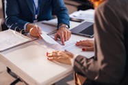 Person in suit pointing to piece of paper across a desk from person with red finger nails