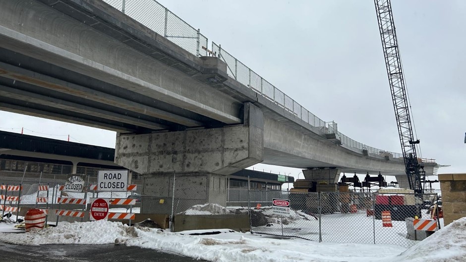 A bridge under construction over a roadway, with a crane on the right side. The area is blocked off by fencing and “Road closed” signs.