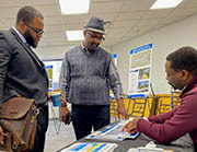 Man at table shows design map to two standing men.