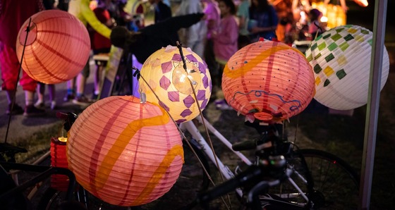 Colorful illuminated paper lanterns decorated with nature-inspired patterns hang among bicycles during an evening outdoor gathering.