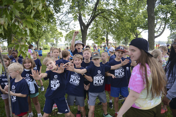 A group of RecPlus kids in navy blue t-shirts rambunctiously pose for a photo at an outdoor picnic