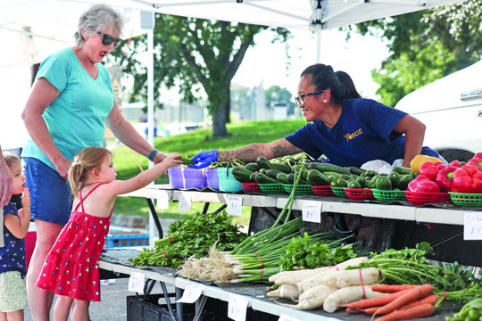 Photo of young girl in front of a table of vegetables reaching out to a woman behind the table handing her a cucumber