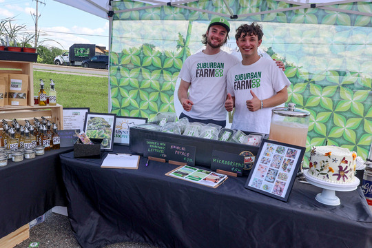 Two young men smiling for the camera behind a table of products and in front of a banner that says Suburban Farm CoOp