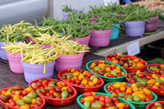 Photo of colorful tomatoes in small baskets and green beans in buckets behind the tomatoes.