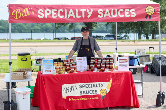 Photo of a man standing behind a table with stacked jars of sauce.