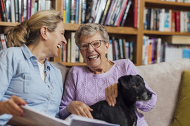 Two adults laughing together looking at a photo album with dog in lap and bookshelf with books in background
