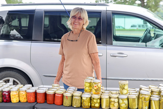 Photo of a woman smiling and standing behind a table with colorful jars of pickles and sauces on it.