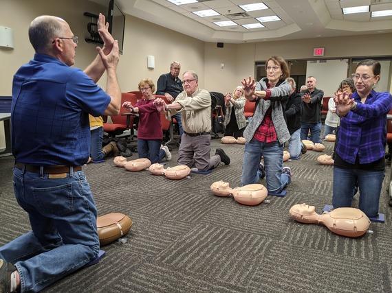 An instructor leads a CPR training course. The class kneels in front of their CPR manikins to perform CPR. 
