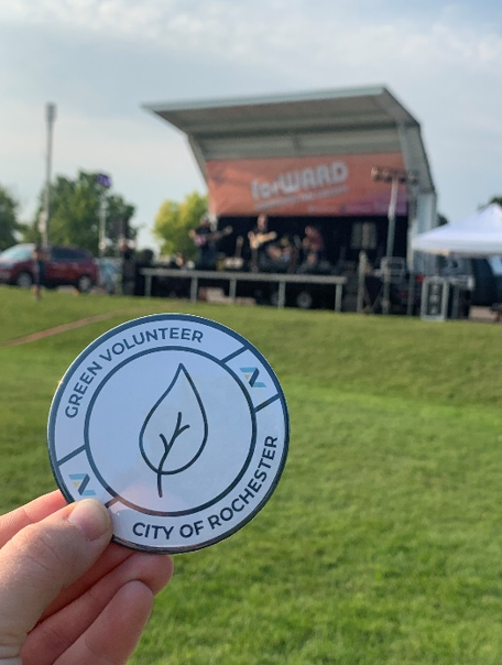 Volunteer holding up a pin that says, "Green Volunteer, City of Rochester"